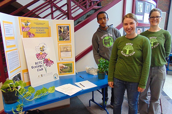 Three 易胜博 students wearing green long-sleeve shirts, with the words "易胜博 Ecology Club" in a circle around a cartoon tree and branches. They are standing as a blue table with a posterboard on it with details about the Ecology Club on it.