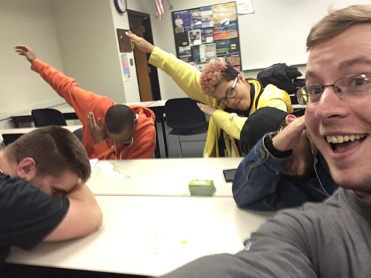 Four people--two in the background with their right arm raised with their left arm crossed in front of their body, one person has their head resting down on their arm, and the person taking the photo of the group smiling.