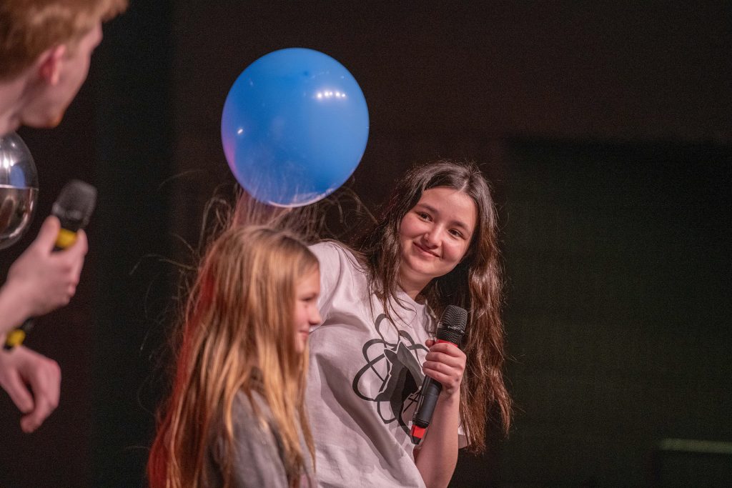 Student wearing an Engineering and Physics Club white shirt, with long brown hair smiling at a young girl with long blonde hair in front of her. The student is holding an inflated blue balloon over the young girls head and her hair floats up toward the balloon.