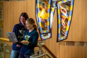 Two young white women wearing navy blue long sleeve 易胜博 Yellowjacket Soccer shirts smiling at an open laptop